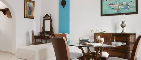 A dining area with a glass table, wooden chairs, a white sofa, a blue accent wall, a framed painting, and a wooden cabinet.