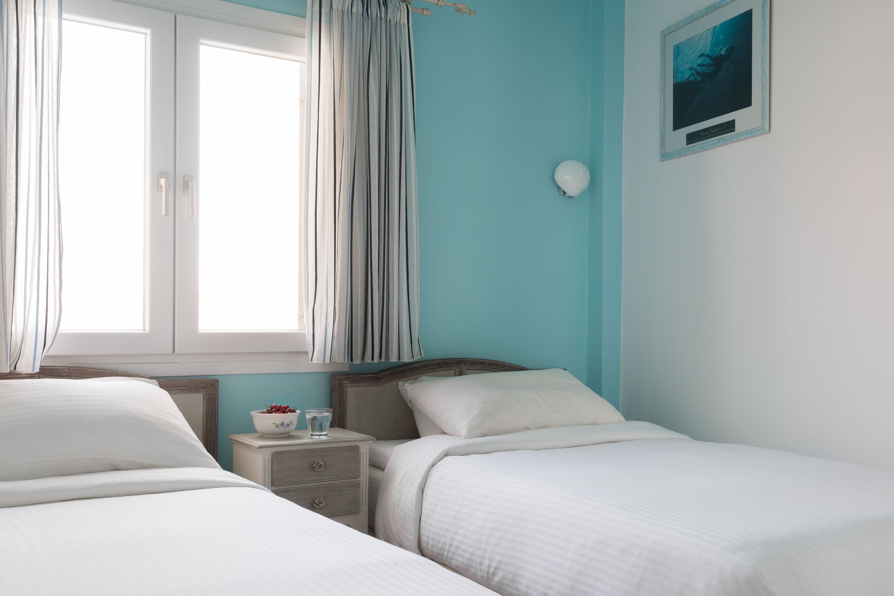 A bedroom with two beds, a nightstand with a bowl of fruit, a bedside table, and a framed picture on the wall.