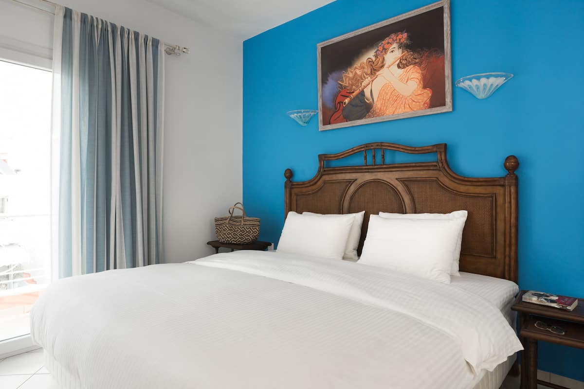 A bedroom with a large bed, a wooden headboard, a painting, a blue wall, and a window with curtains.