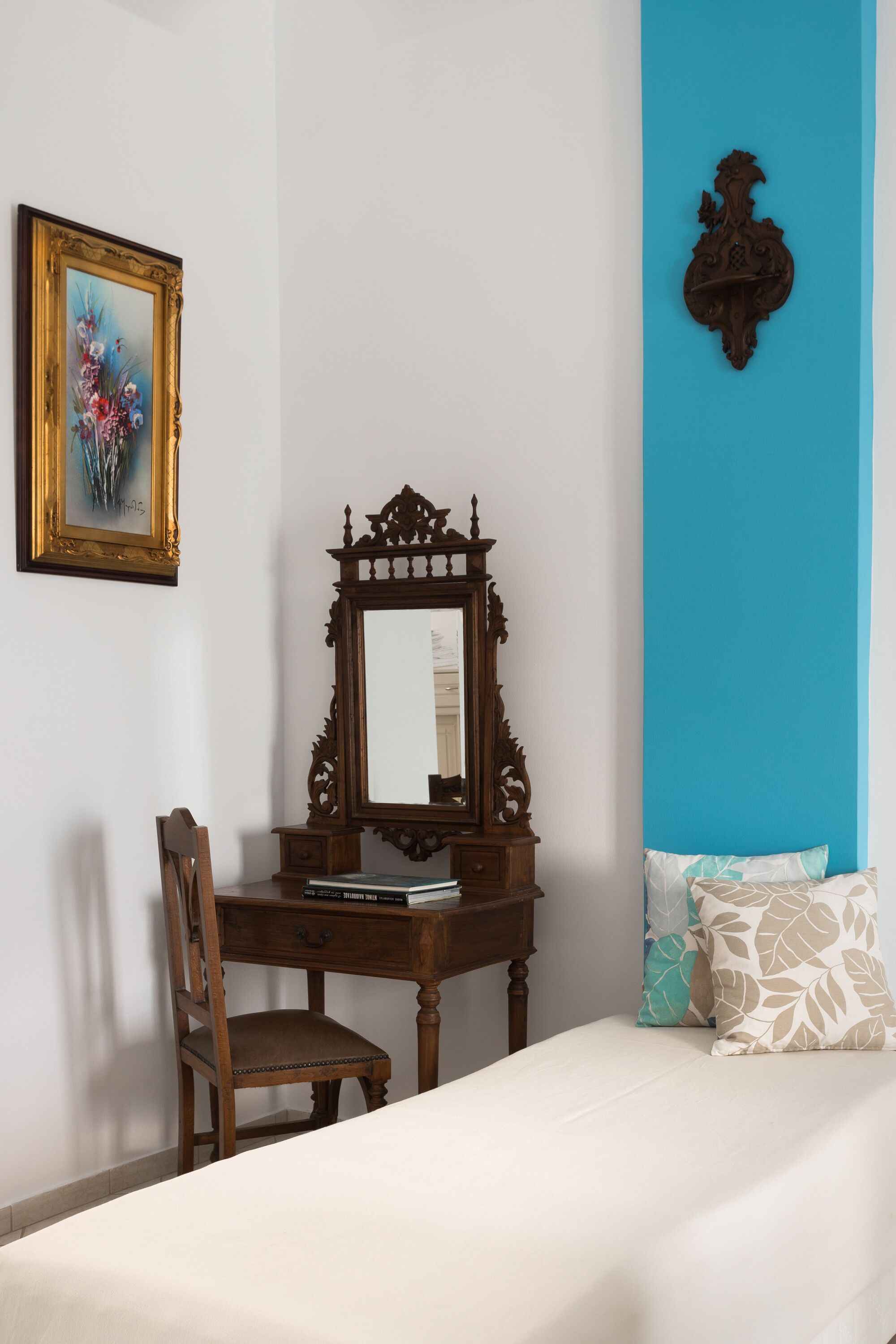 Une chambre avec un bureau en bois, un miroir, un lit agrémenté d’oreillers à motifs et un tableau encadré accroché au mur.