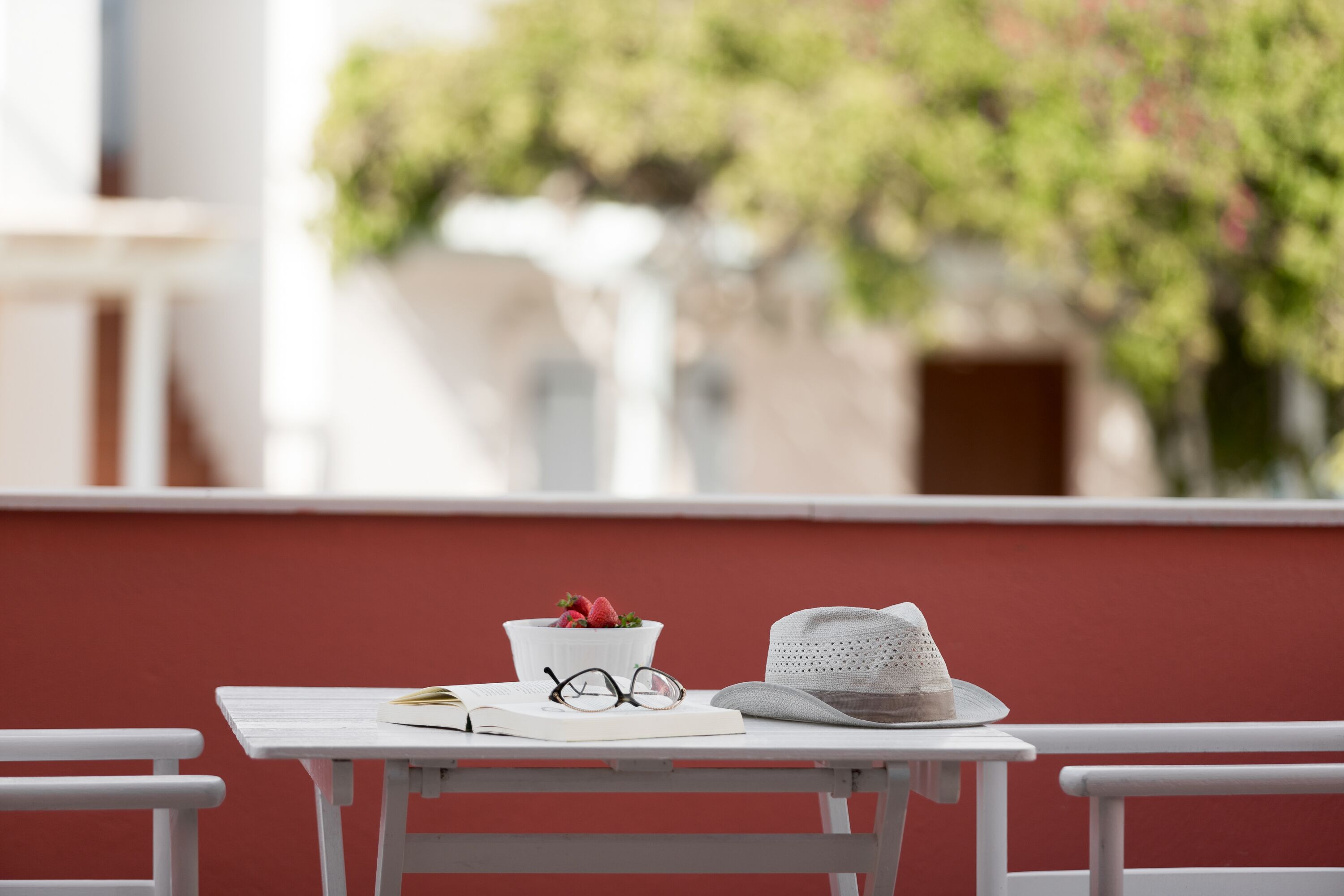 A white table with a book, glasses, a hat, and a bowl with strawberries on a balcony.