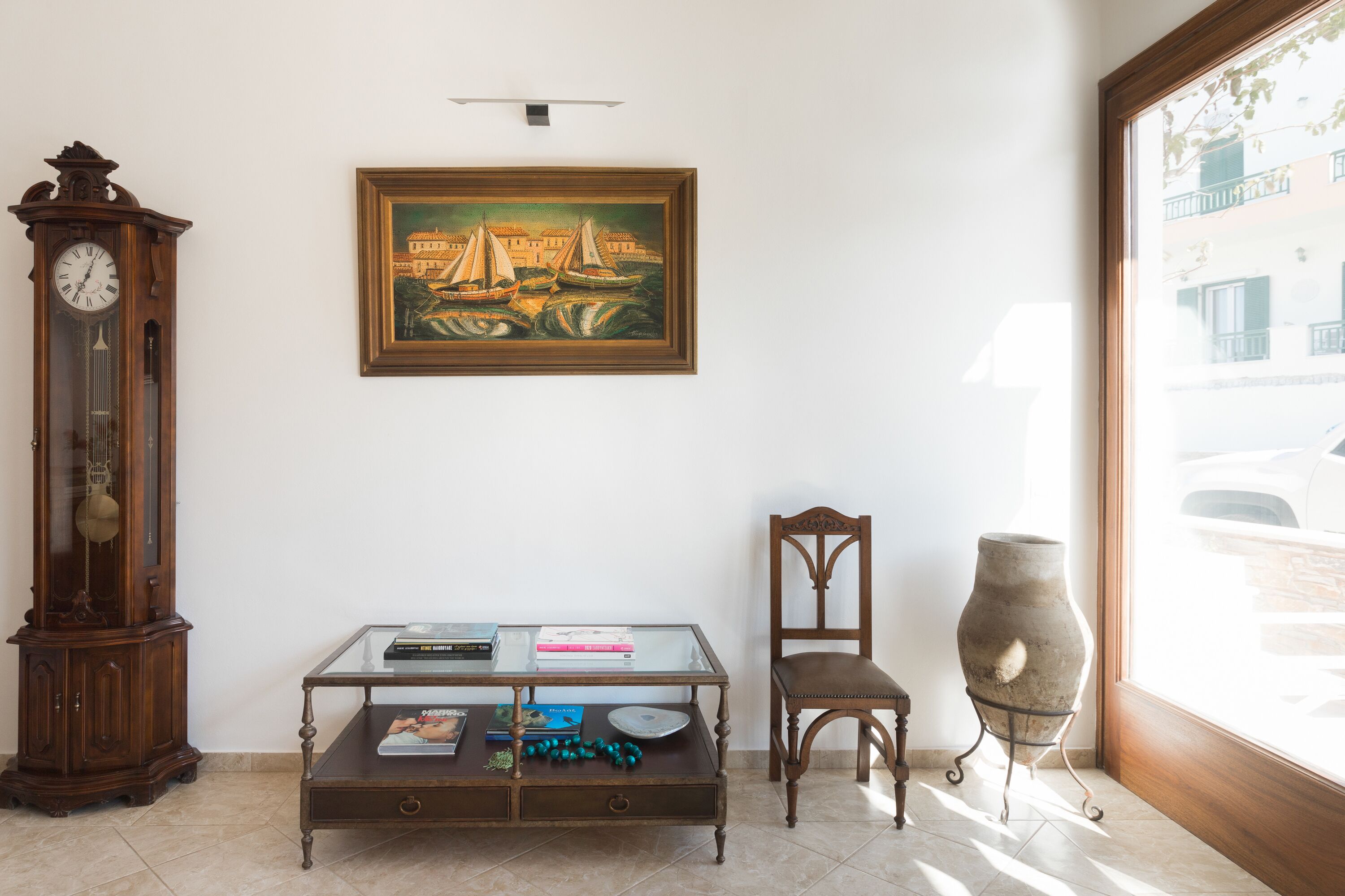 A room with a wooden grandfather clock, a glass-top coffee table with books and decorative items, a framed painting above the table, a wooden chair, and a large window with a view of a building outside.