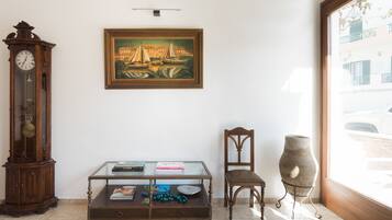 A room with a wooden grandfather clock, a glass-top coffee table with books and decorative items, a framed painting above the table, a wooden chair, and a large window with a view of a building outside.