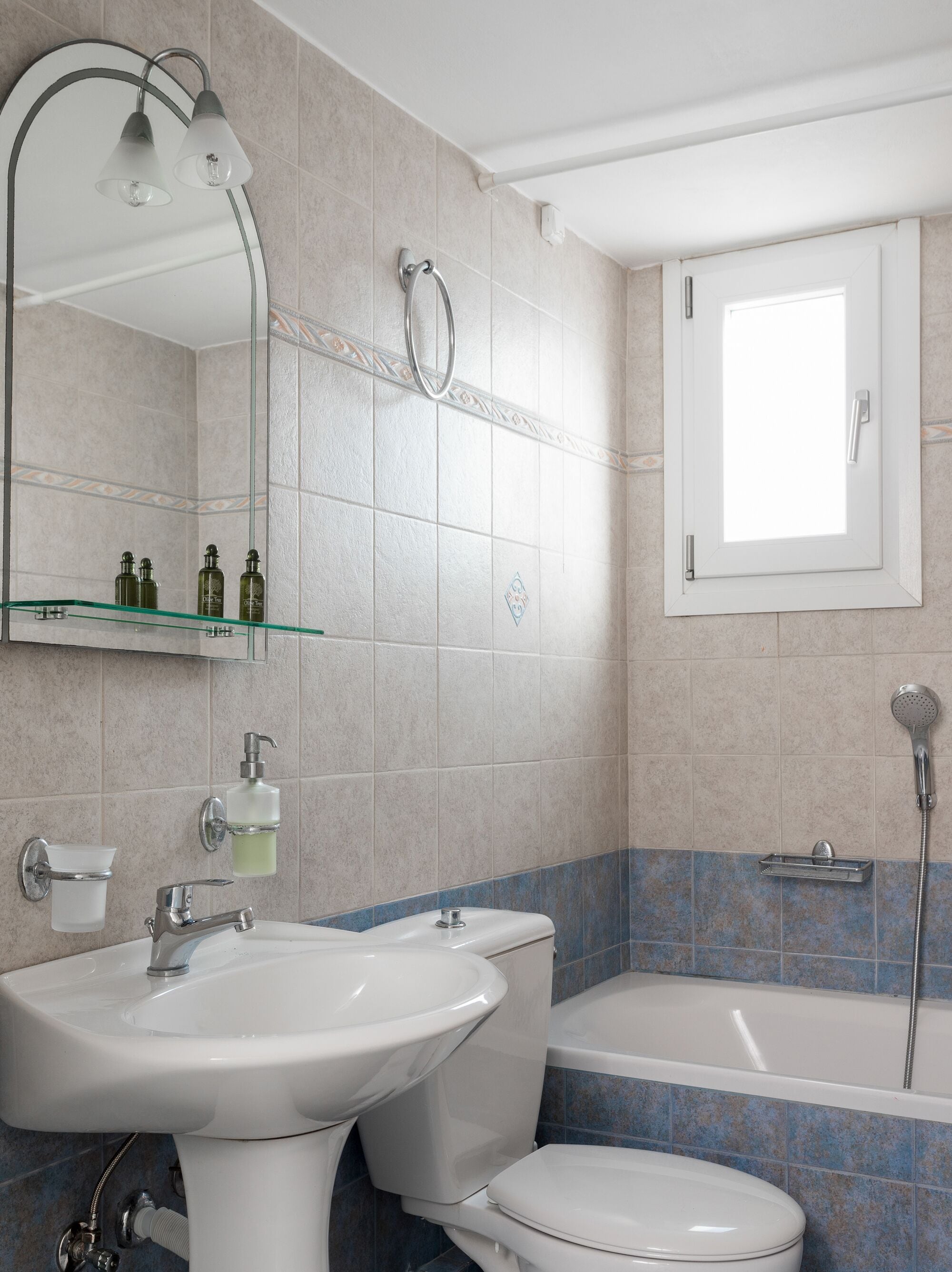 Een badkamer met een wastafel, toilet en bad. Er is een spiegel met verlichting, een douchekop en een raam.