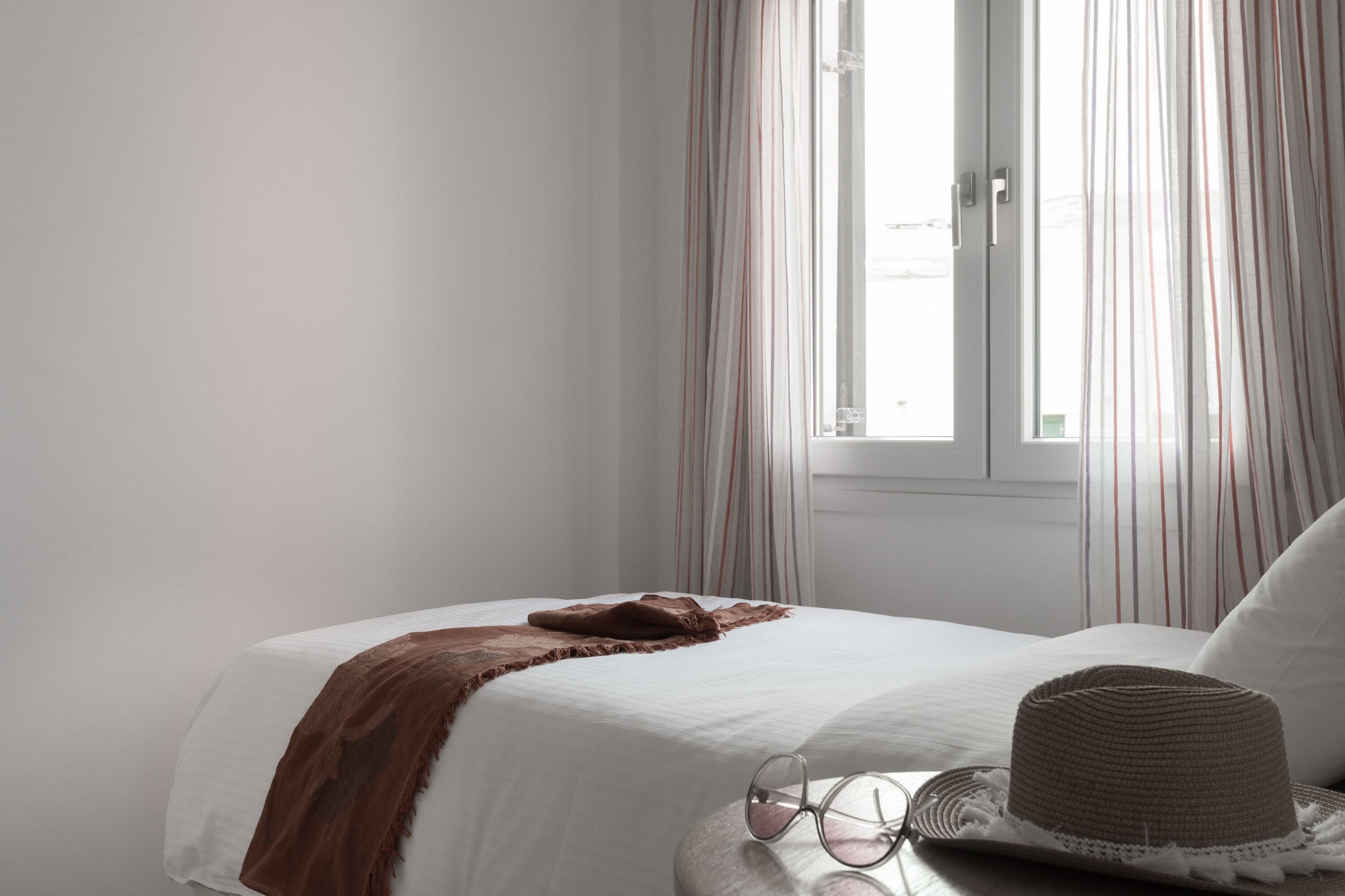 Una cama bien hecha con sábana blanca y manta marrón, un sombrero y gafas sobre una mesita de noche.