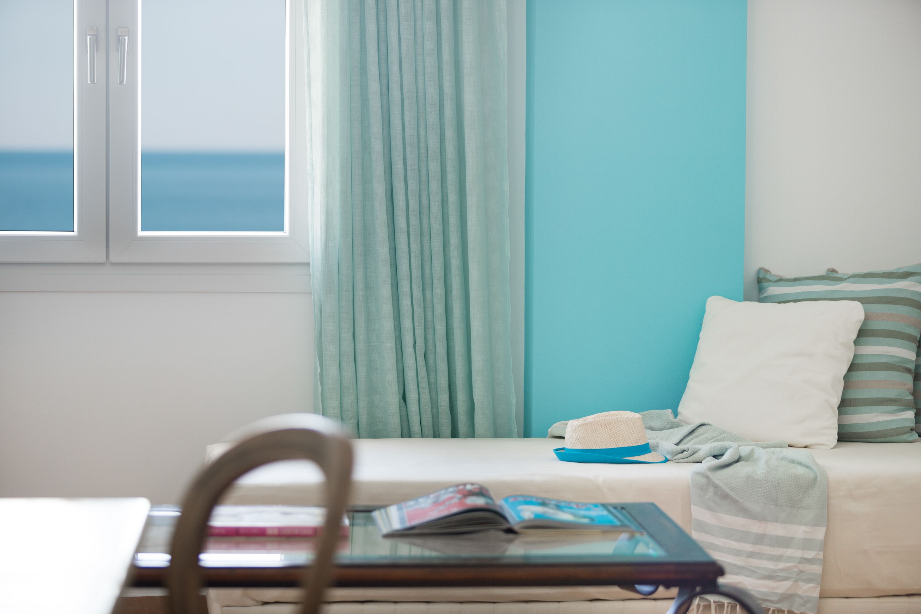 A room with a bed, a window with curtains, a table with a magazine, and a chair.