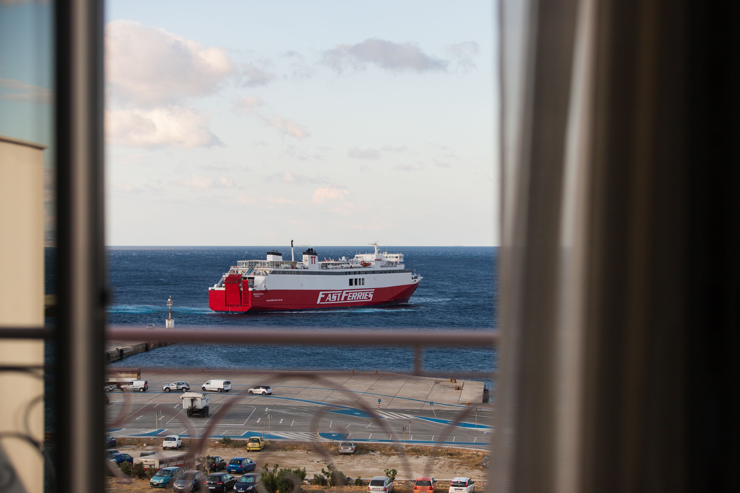 Un ferry llamado 'FAST FERRIES' pasa por una carretera costera con coches aparcados y un cielo despejado.