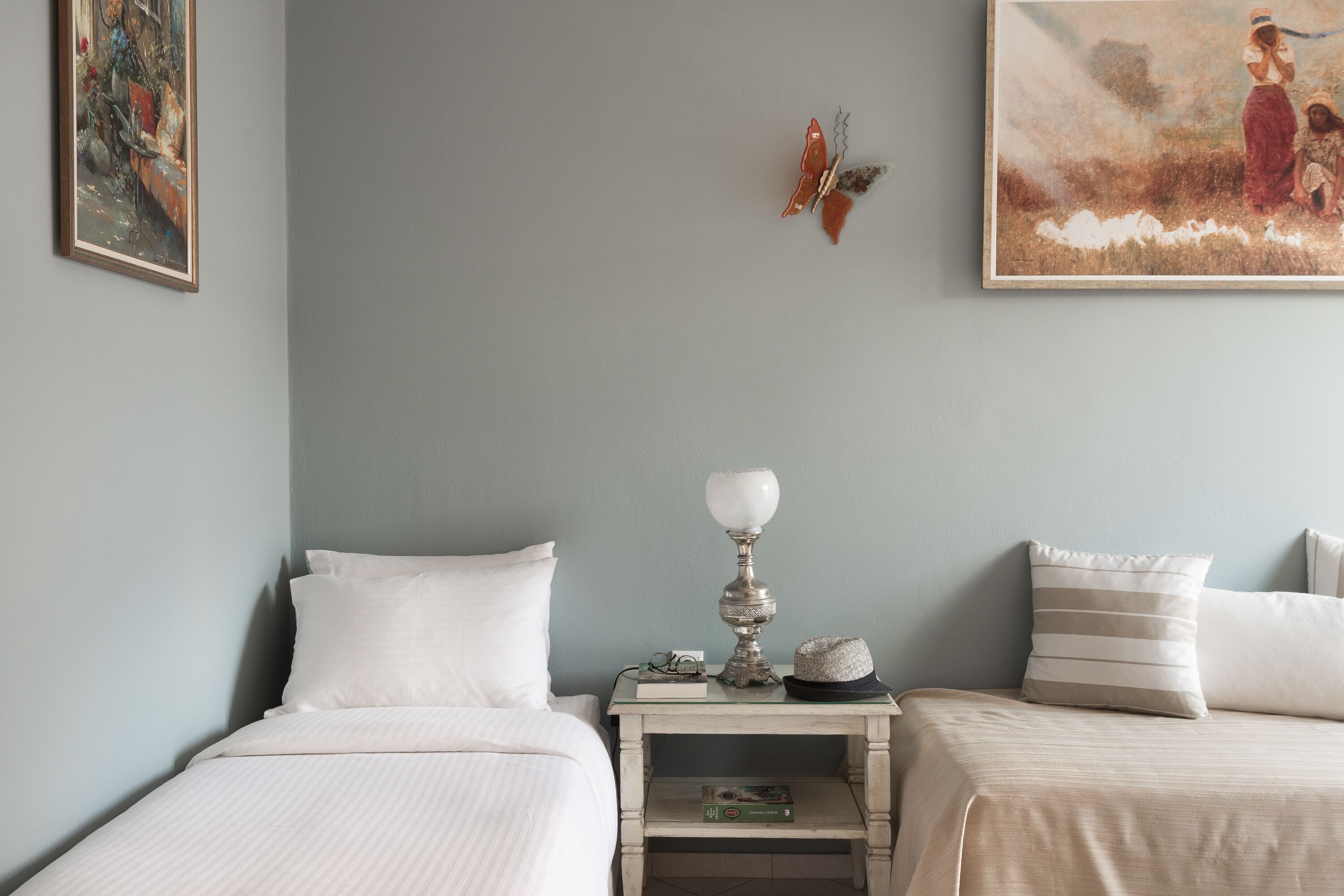A bedroom with two beds, a nightstand with a lamp, a book, and a hat. There are paintings on the walls.