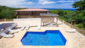 Une piscine avec des chaises longues, un espace salon couvert et une vue sur une maison avec un toit rouge.