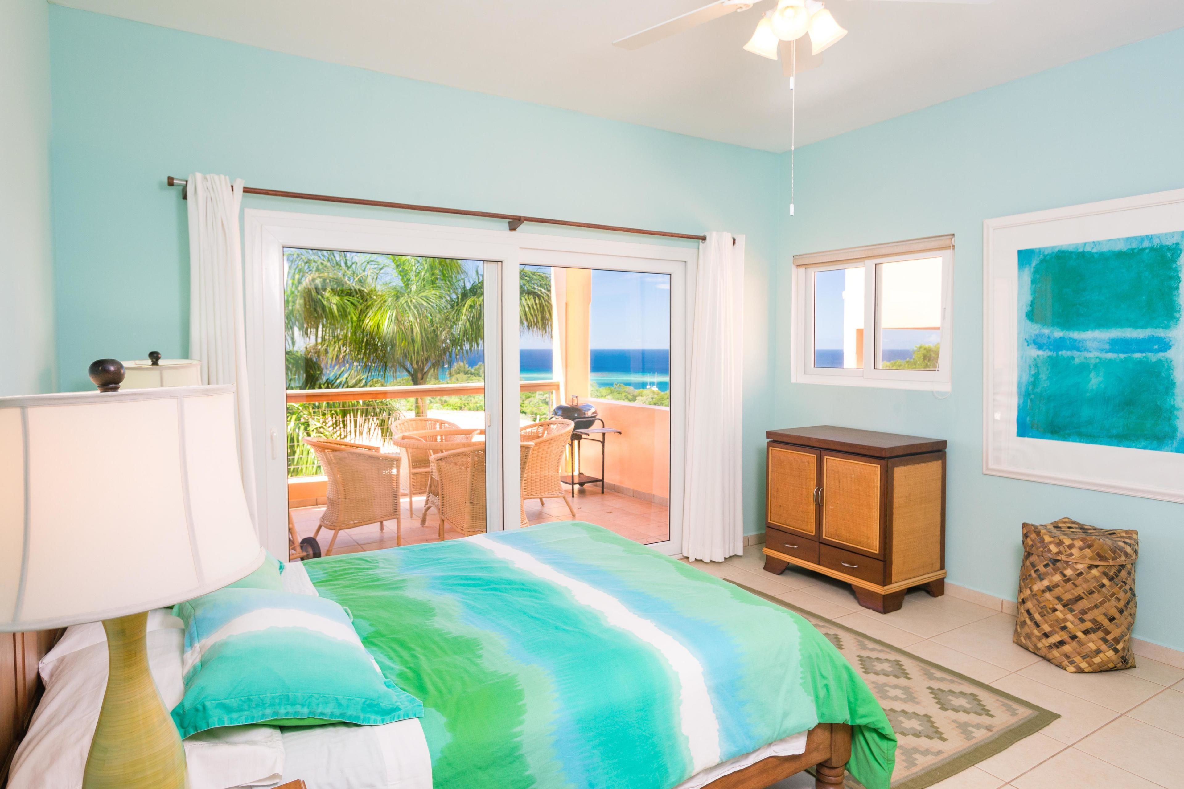 A bedroom with a bed, a nightstand, a lamp, a window, and a view of the outdoors.