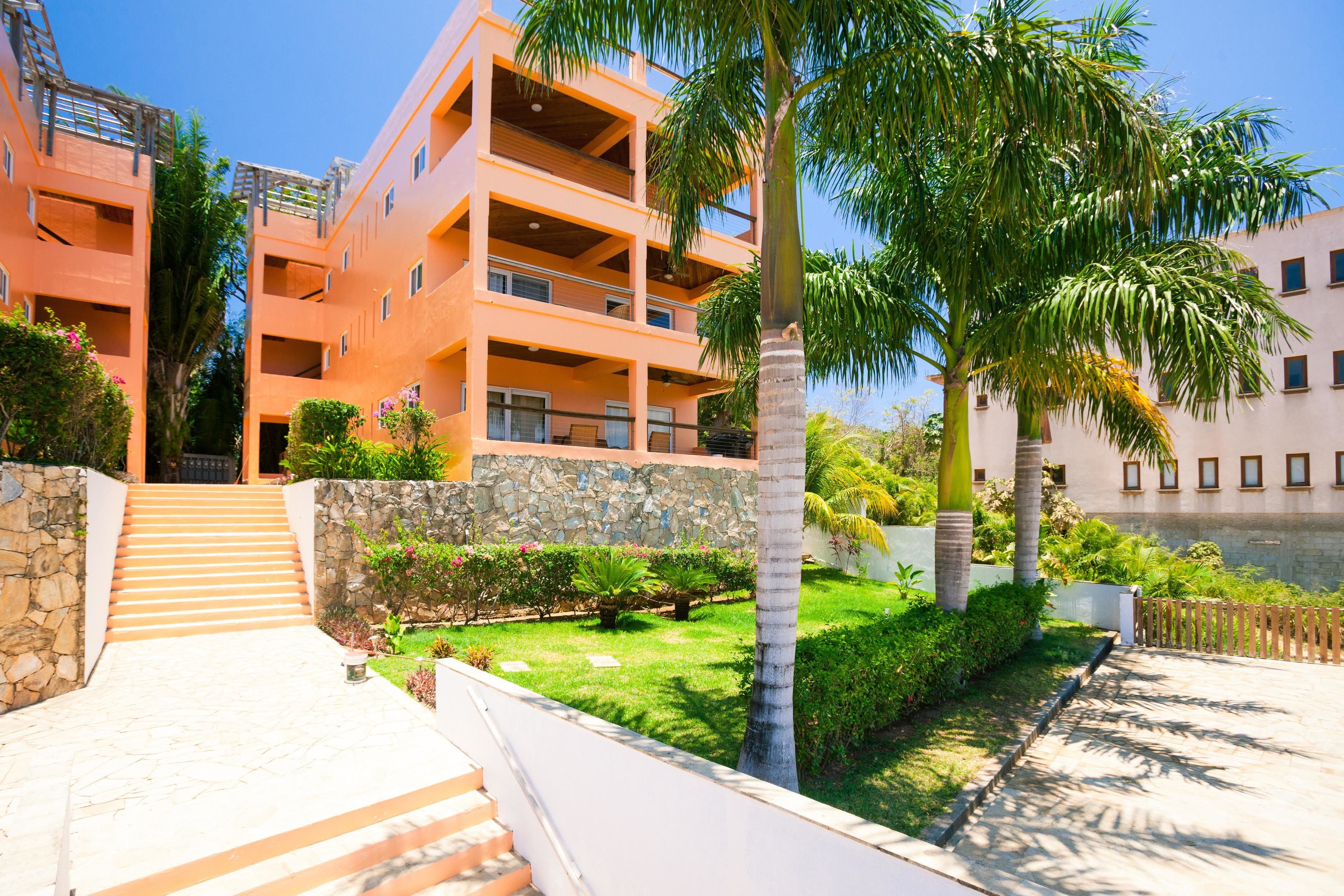 A multi-story building with balconies, a stone wall, and a well-maintained garden with palm trees.