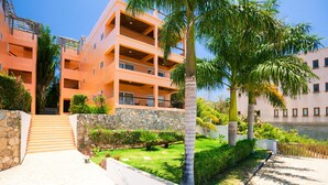 A multi-story building with balconies, a stone wall, and a well-maintained garden with palm trees.