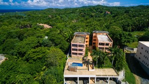 A resort with multiple buildings surrounded by lush greenery, a swimming pool, and a view of the ocean.