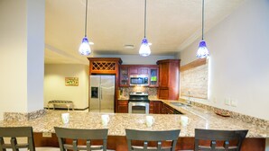 A kitchen with granite countertops, wooden cabinets, a refrigerator, microwave, and a stove. There are three bar stools and pendant lights. - Villa Del Playa 3 (Roatan)