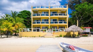 A multi-story yellow building with balconies, a beach in the foreground, and a boat docked on the sand. - Villa Del Playa 3 (Roatan)