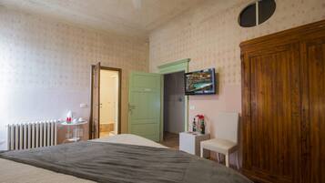 A bedroom with a bed, a wooden wardrobe, a small table with a chair, a radiator, and a door leading to another room.