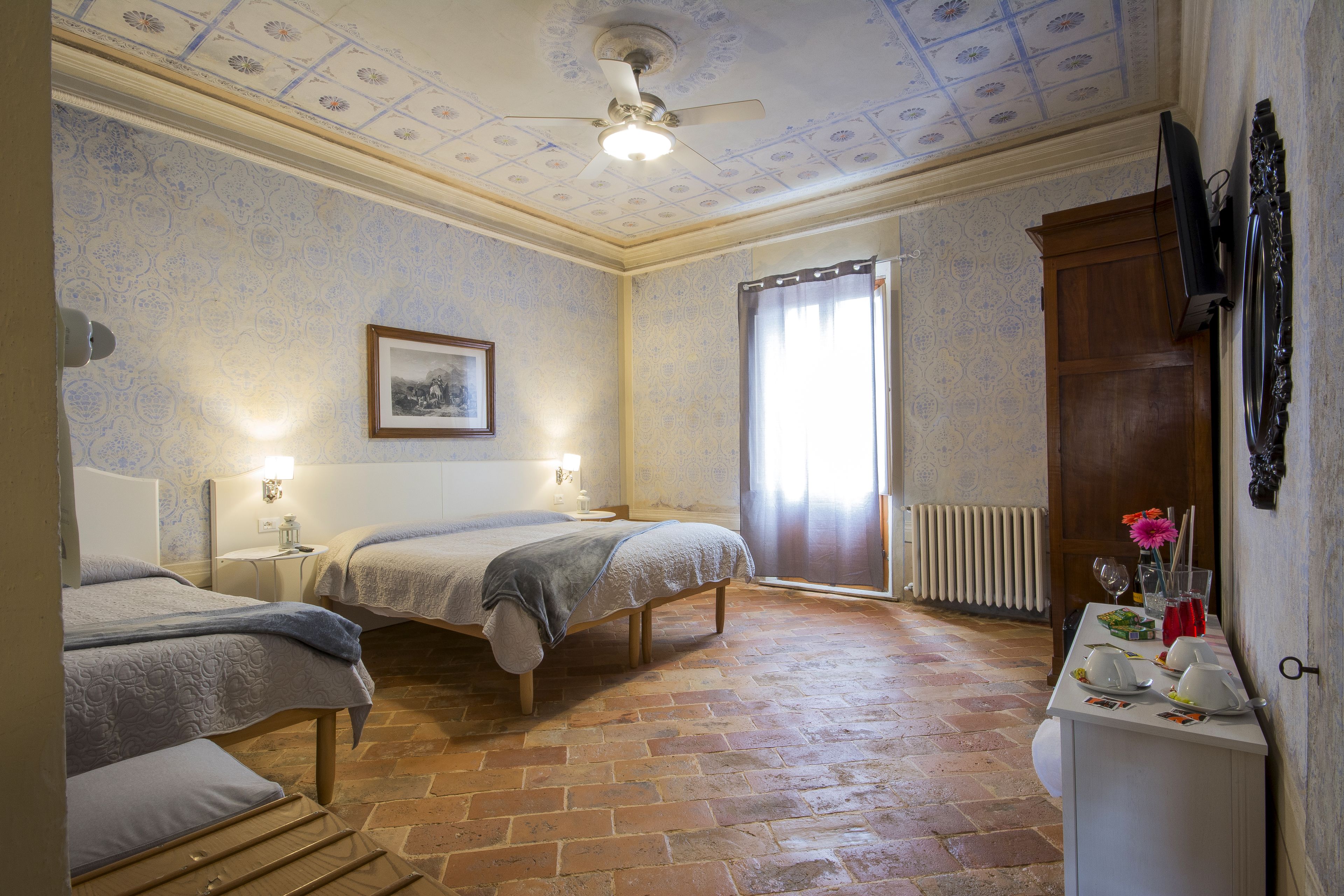 A room with two beds, a ceiling fan, a framed picture on the wall, a radiator, and a table with items on it.