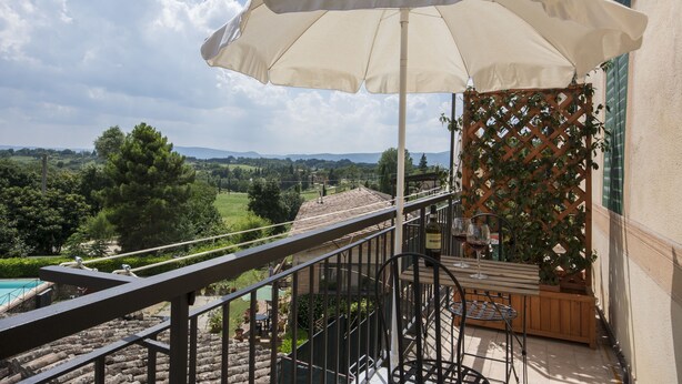 A balcony with a view of a pool and landscape, a wooden table with chairs, and a large umbrella. - Villa Sant Andrea (Siena)