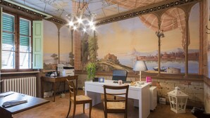 A room with a desk, chairs, a radiator, a window with shutters, a mural, and a chandelier. - Villa Sant Andrea (Siena)