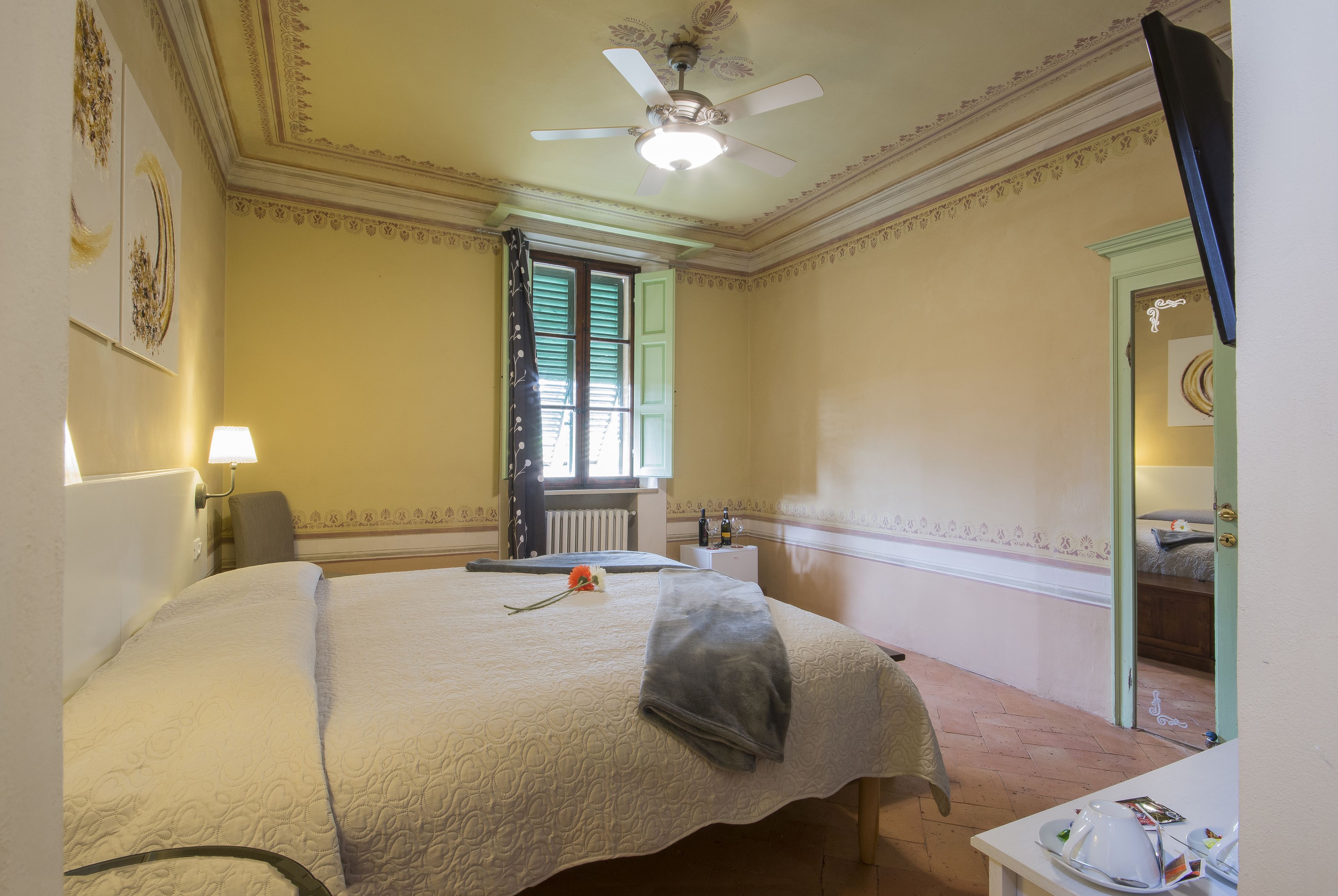 Hotelkamer met een groot bed, een plafondventilator, een raam met luiken, een radiator en een kleine tafel met een lamp.