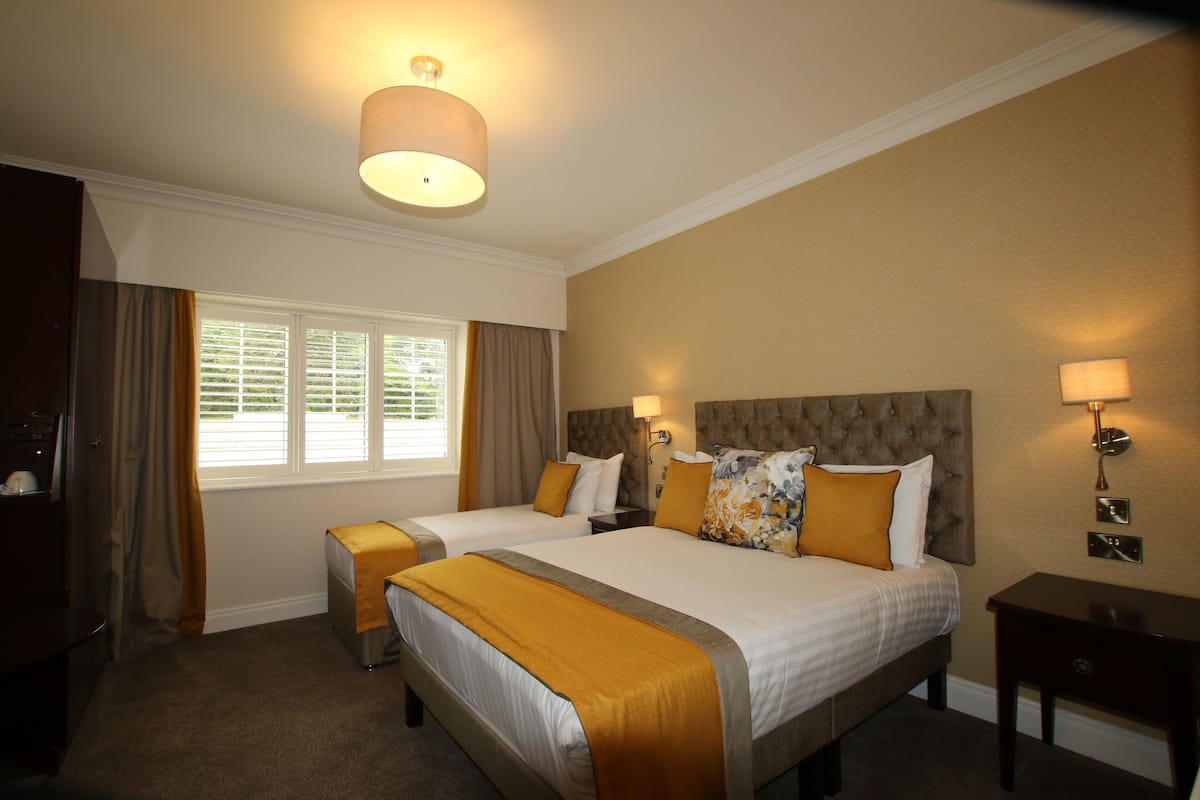 A hotel room with two beds, a window with blinds, a bedside table, and a lamp.