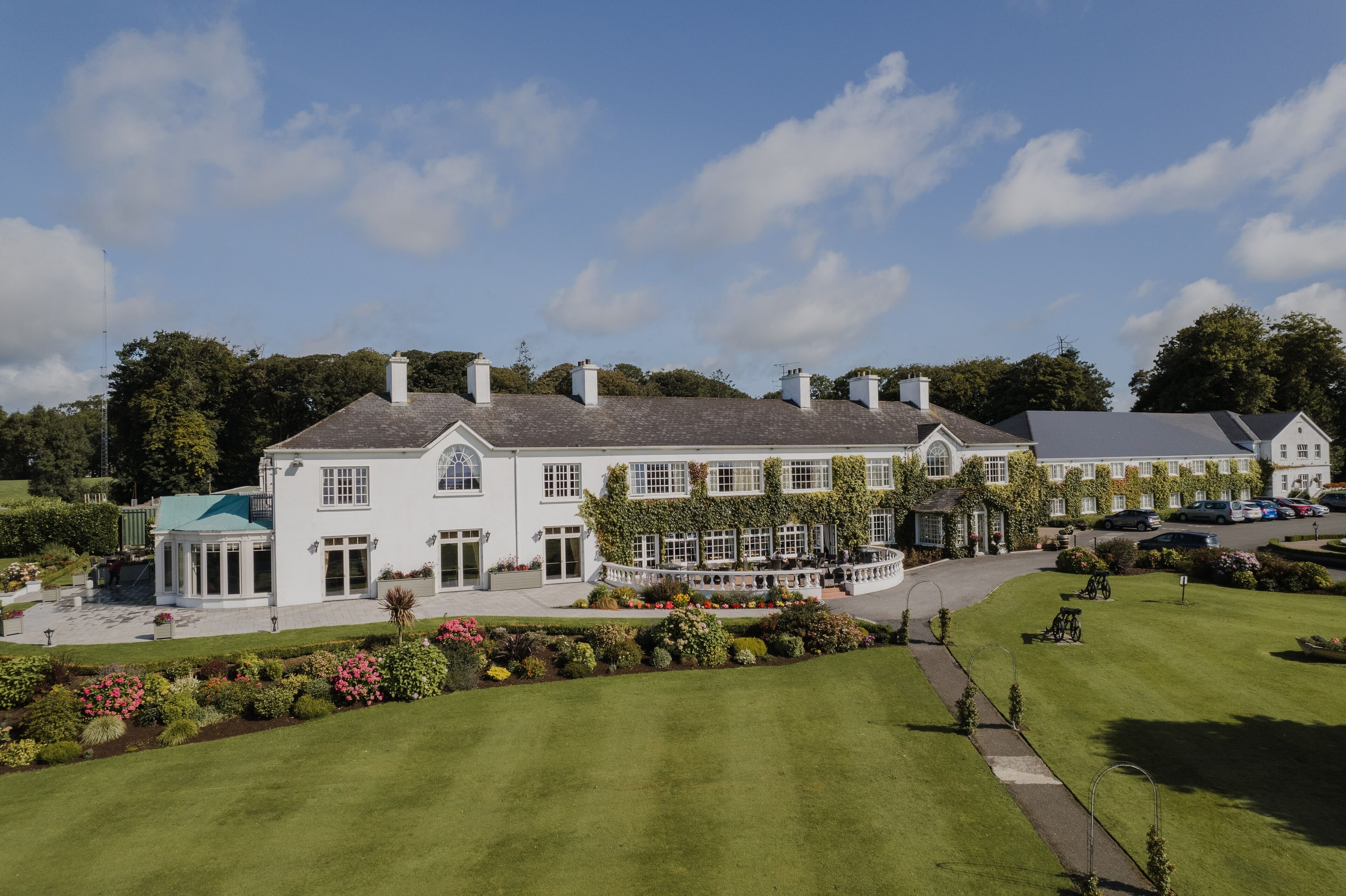A large, white, two-story building with a green roof, surrounded by well-maintained gardens and a clear sky.