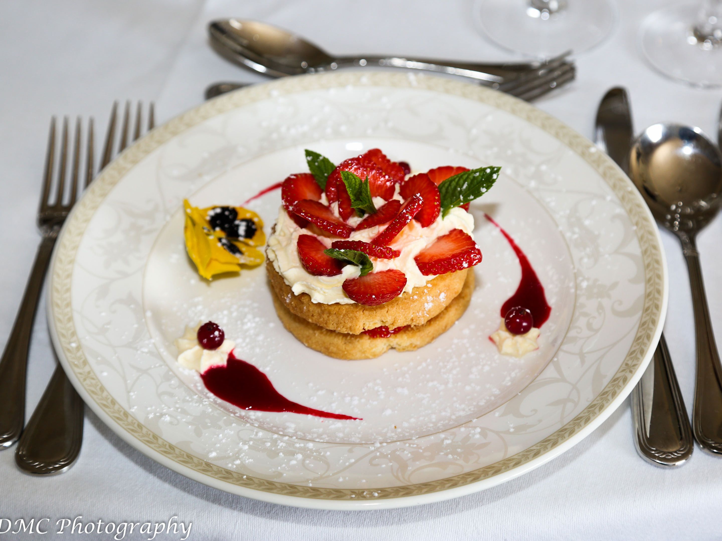 Een dessert op een wit bord met een gouden rand, versierd met aardbeien en muntblaadjes, en een bessen-saus.