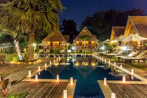 Un resort nocturno con piscina central, terraza de madera y villas con techo de paja.