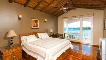 Quarto com teto de madeira, cama com colcha branca, duas luminárias de cabeceira, mesa de cabeceira, cômoda de madeira e televisão.