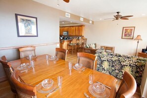 Comedor con mesa de madera dispuesta para seis personas, cocina al fondo y sofá estampado.