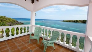 Un balcón techado con dos sillas Adirondack verdes, piso de baldosas y vista al mar y la costa distante.