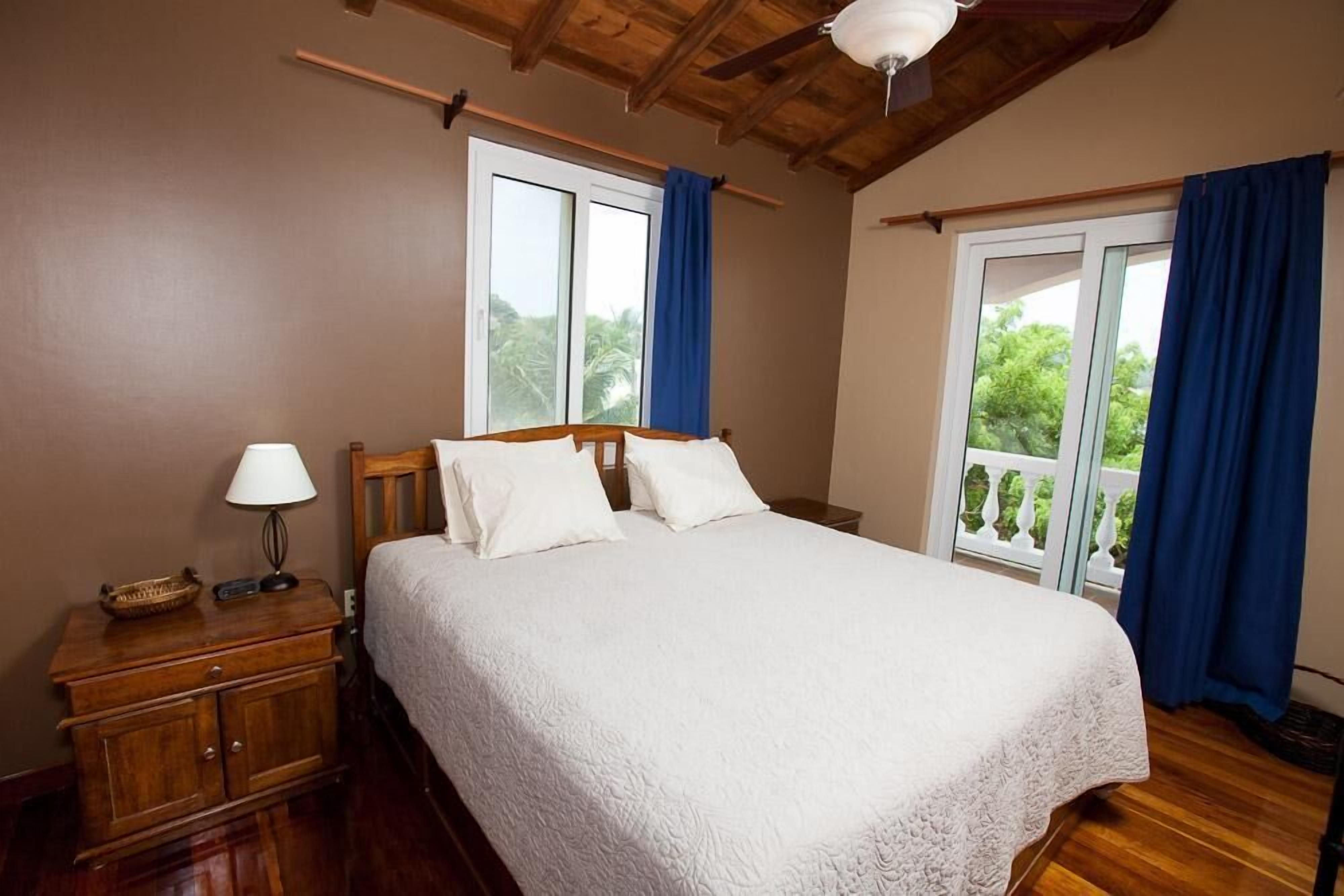 A bedroom with a wooden bed, a nightstand, a lamp, and a view of greenery through the window.