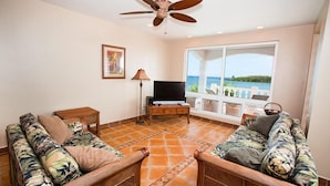 A living room with wicker furniture, a ceiling fan, a TV, and a large window with a view of the sea.