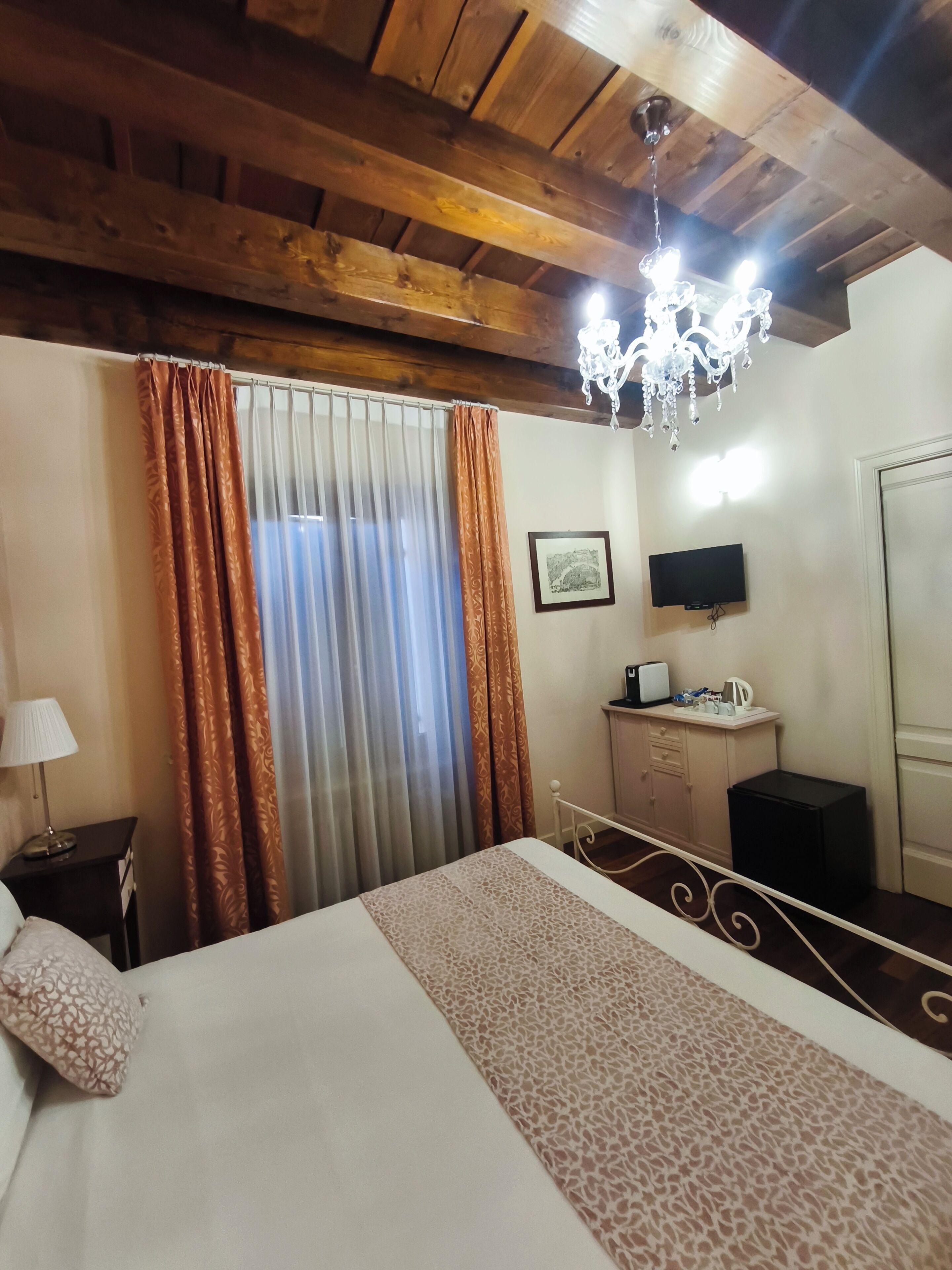 A hotel room with a bed, curtains, a chandelier, a television, a small cabinet, and a desk with a lamp.