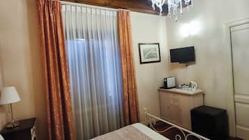 A hotel room with a bed, curtains, a chandelier, a television, a small cabinet, and a desk with a lamp.