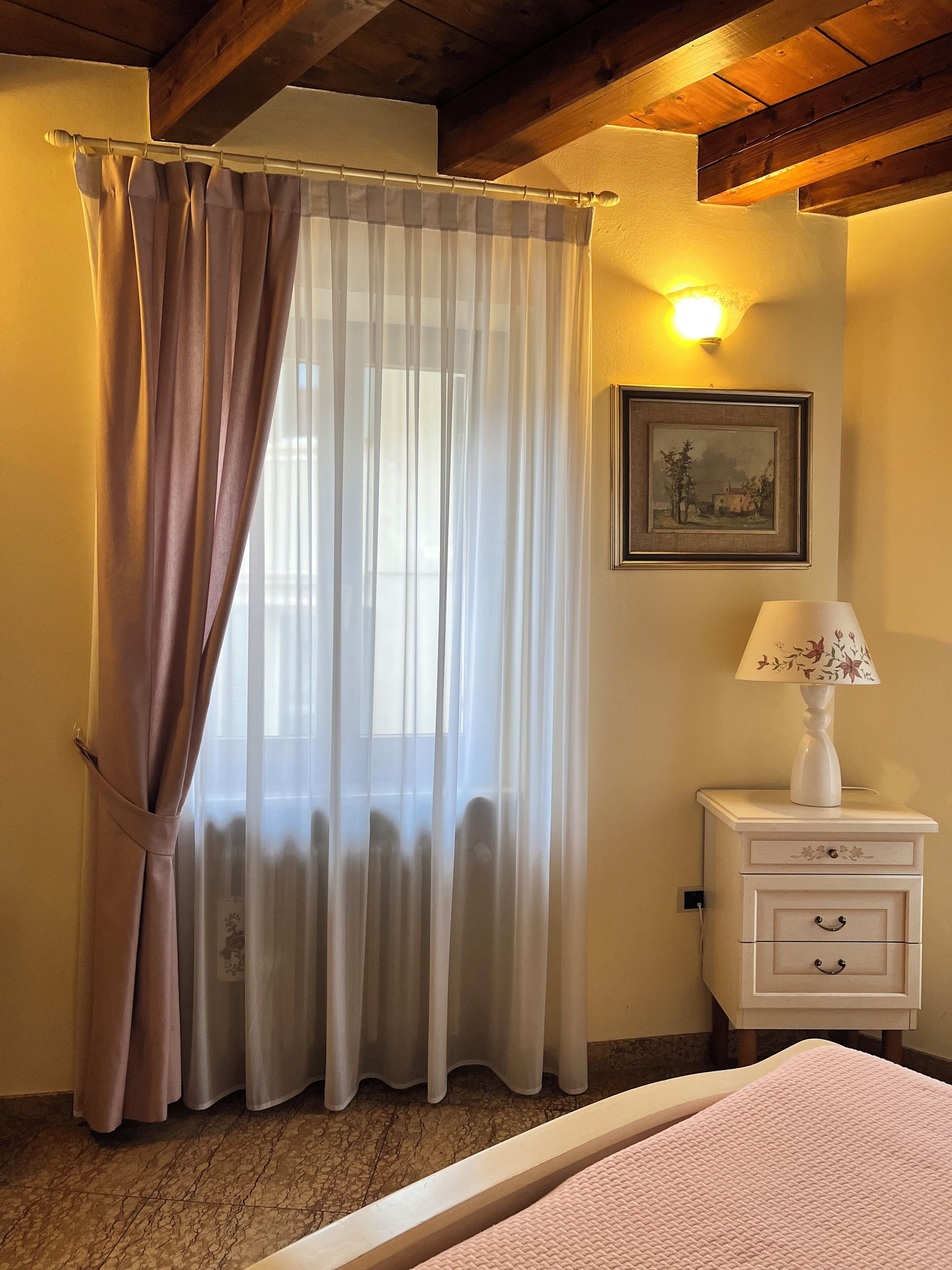 A bedroom with a bed, a nightstand with a lamp, a window with sheer curtains, a framed picture on the wall, and a wooden ceiling.