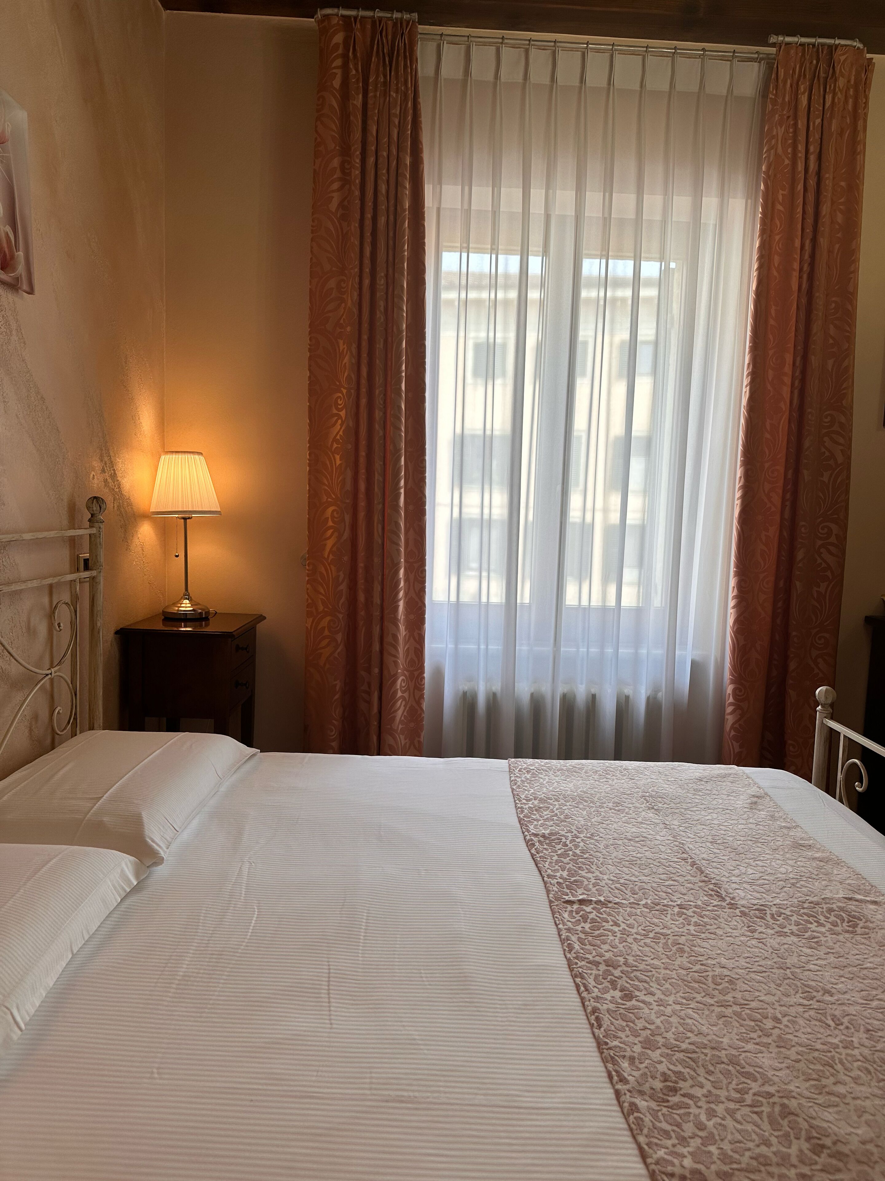 Una habitación de hotel con cama, una mesita de noche con lámpara y una ventana con cortinas translúcidas.