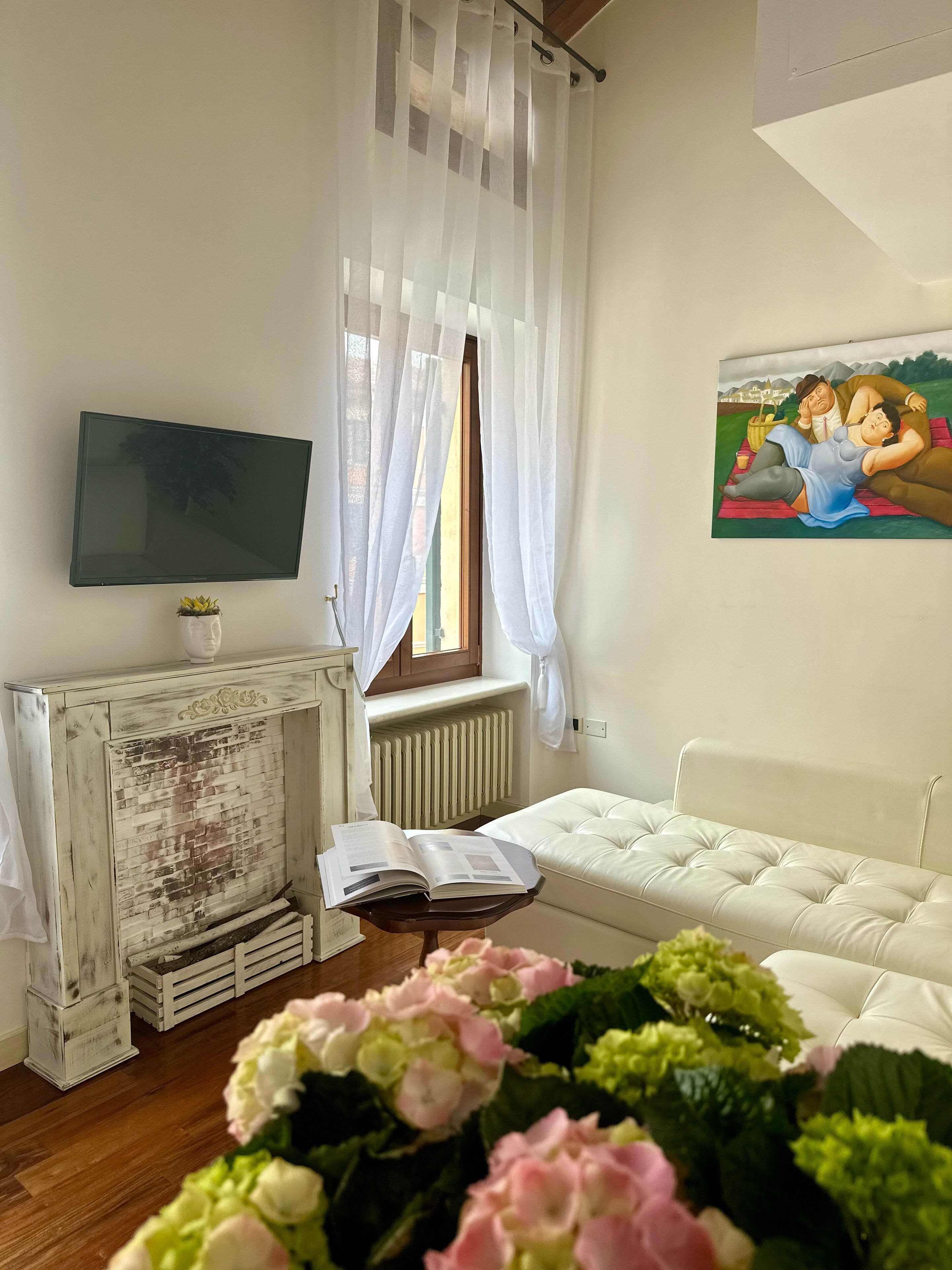 A living room with a white sofa, a flat-screen TV mounted on the wall, a fireplace with a rustic design, and a window with sheer curtains.