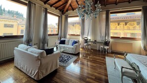 A spacious living room with wooden flooring, a chandelier, a dining area, and large windows offering a view of buildings and trees. - Relais A Ponte Pietra (Verona)