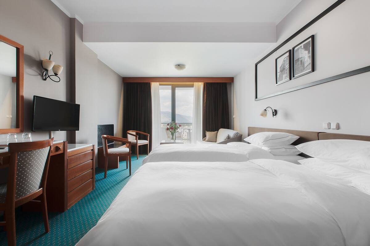 Ein Hotelzimmer mit einem Bett, einem Schreibtisch, einem Stuhl, einem Fernseher und einem fensterbepflanzten Fenster.