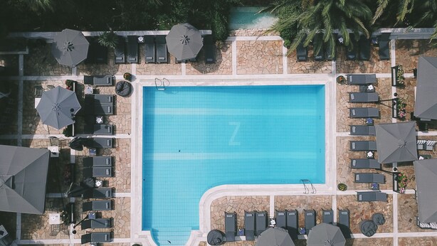 Vista aérea de una piscina rectangular rodeada de sillas de descanso y sombrillas, con muros de piedra y palmeras al fondo.