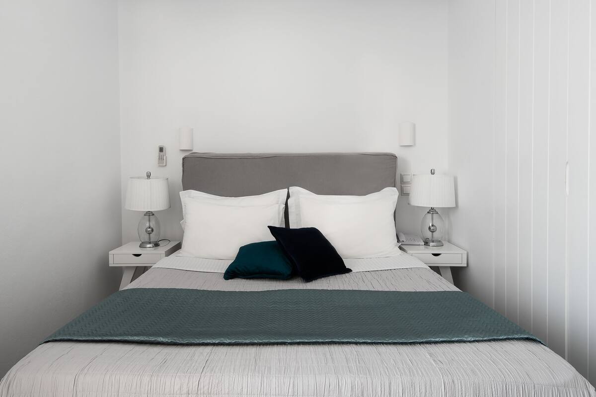 Una cama bien hecha con cabecera gris, ropa de cama blanca y dos lámparas de noche.