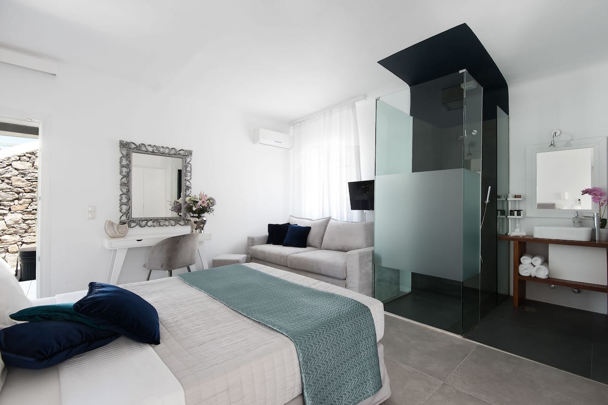 Un dormitorio moderno con cama, sofá, cabina de ducha con paredes de cristal, tocador con espejo y pared de piedra.