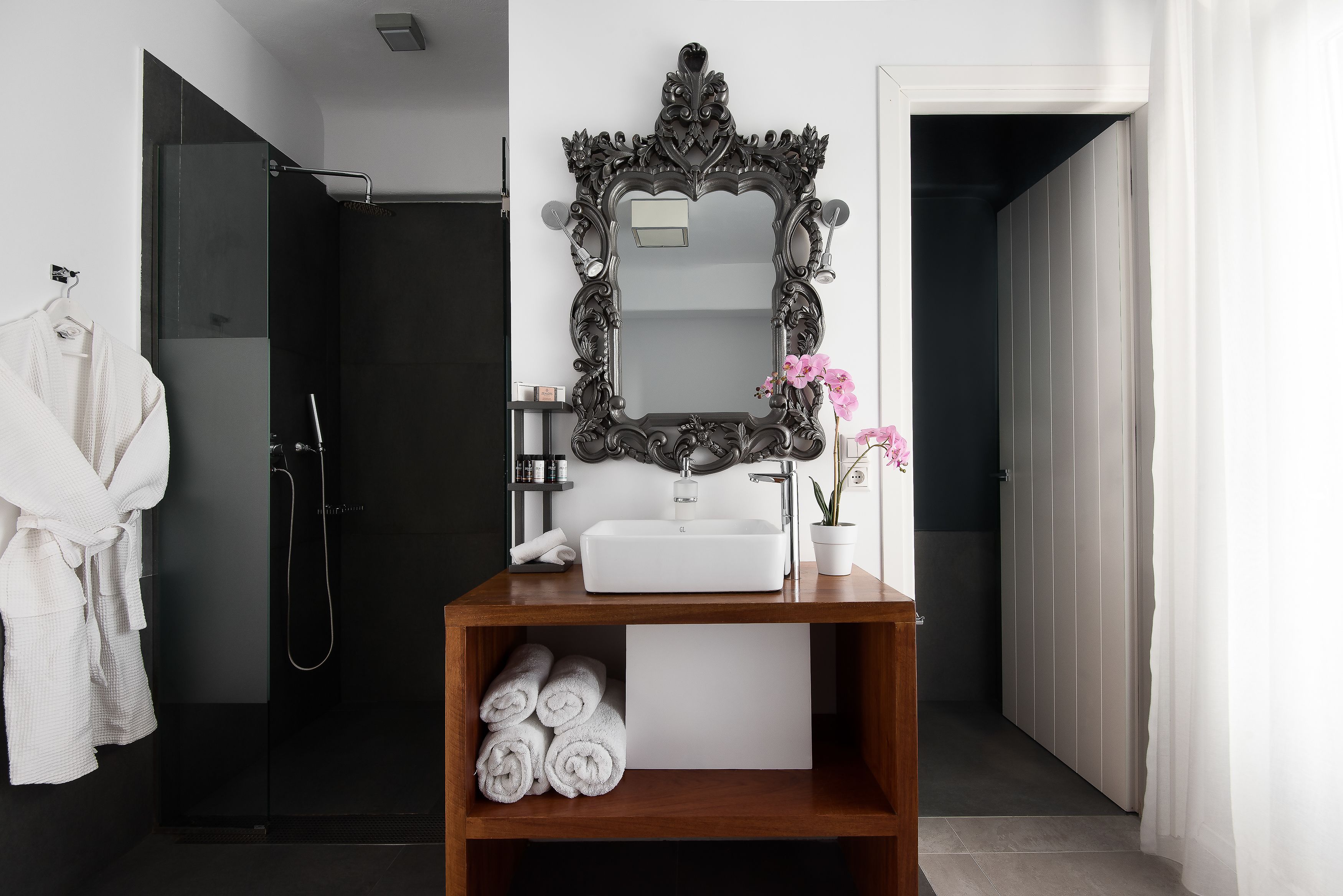 A modern bathroom with a large mirror, white sink, and a wooden shelf with towels.