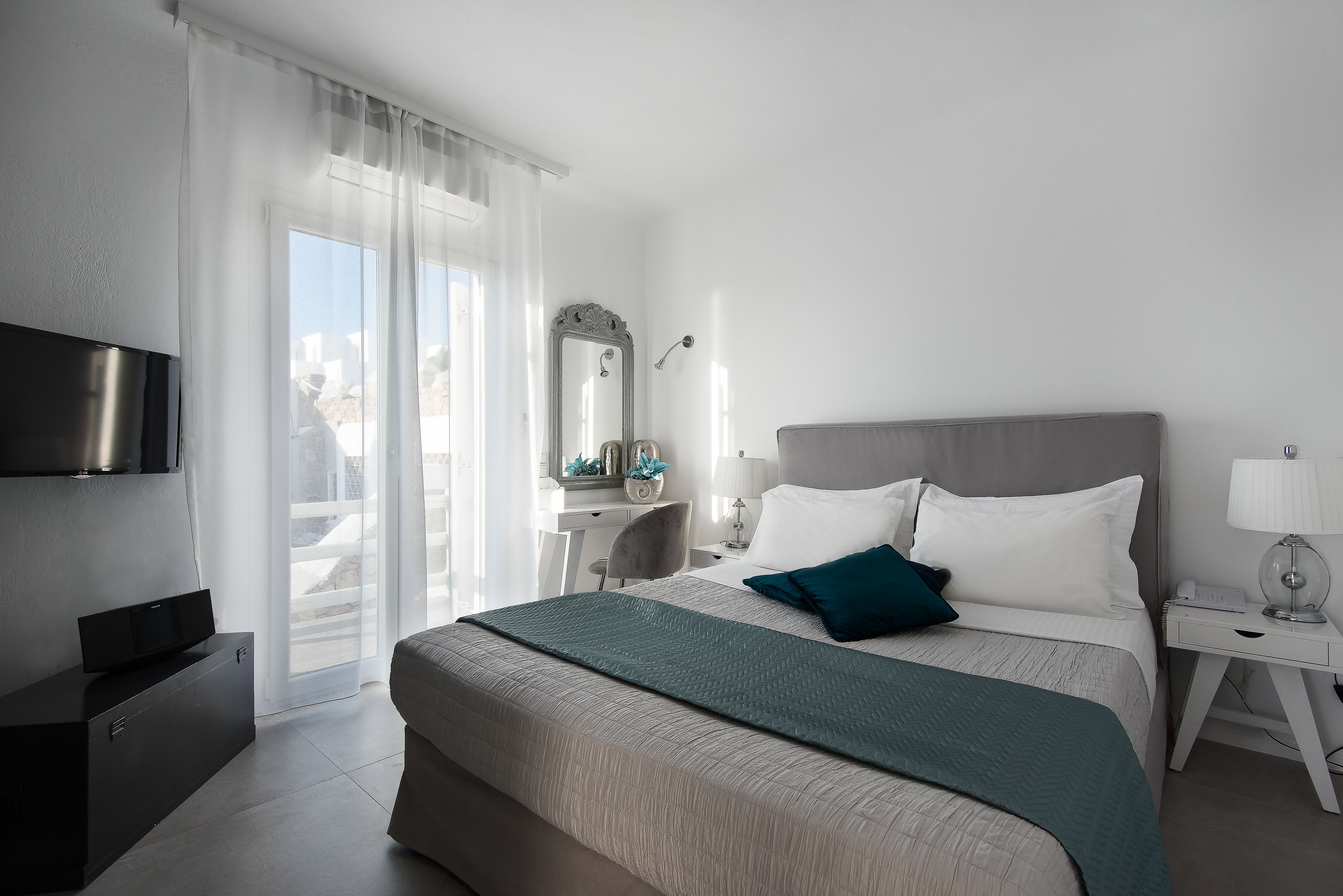A modern hotel room with a large bed, a flat-screen TV, a desk with a chair, a mirror, and a balcony with a view.