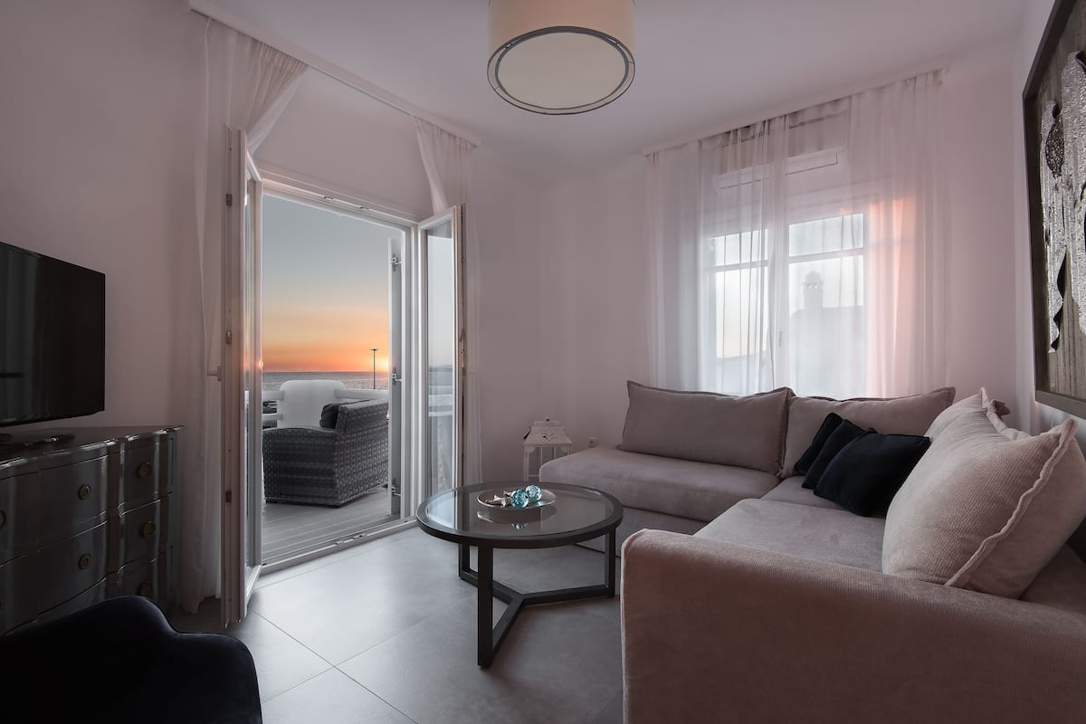 Una sala de estar moderna con sofá, mesa de centro, televisor y vista al atardecer a través de la puerta abierta.