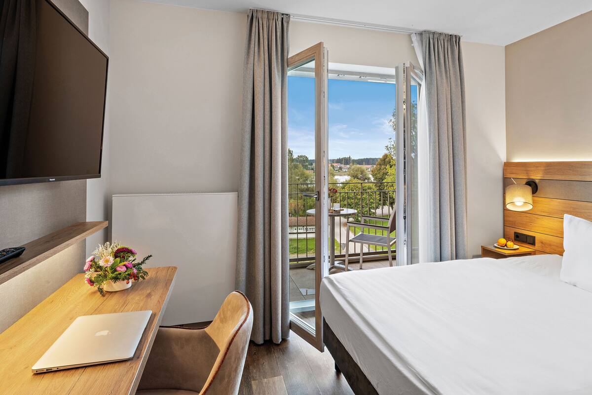 A hotel room with a bed, a desk with a laptop, a TV, and a balcony with a view of the countryside.