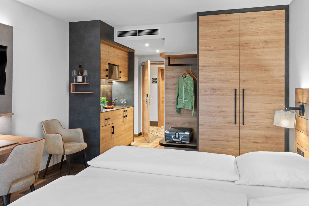 A modern hotel room with a bed, a desk with a laptop, a TV, a wardrobe, and a kitchenette.