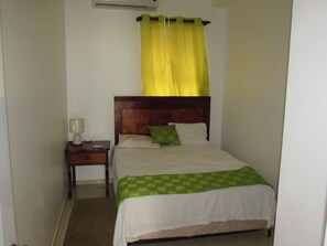 Una cama individual con cabecera de madera, una colcha verde a cuadros y una cortina amarilla. Se ve un mesita de noche con una lámpara y una pequeña mesa con otra lámpara.