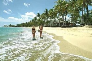 Zwei Personen spazieren am sandigen Strand entlang, im Hintergrund Palmen und das Meer.